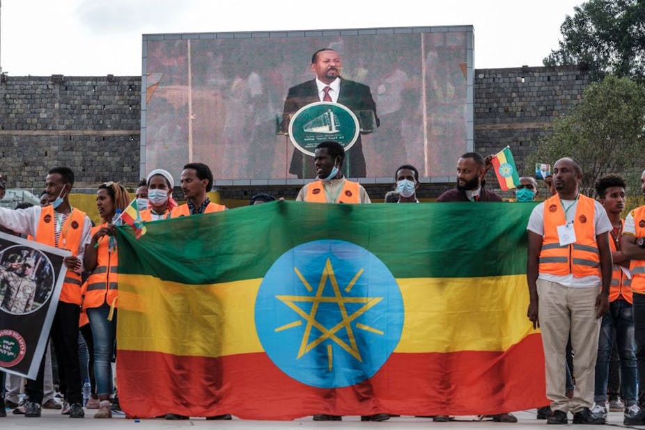A man's image is projected on a large TV screen with a crowd standing in front of it holding up a flag with green, yellow and red stripes, with a blue circle in the middle and a yellow star