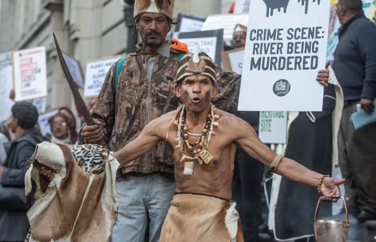 Protesters holding placards outside a court, a man in front spreads his arms, bare-chested and wearing traditional animal skins.