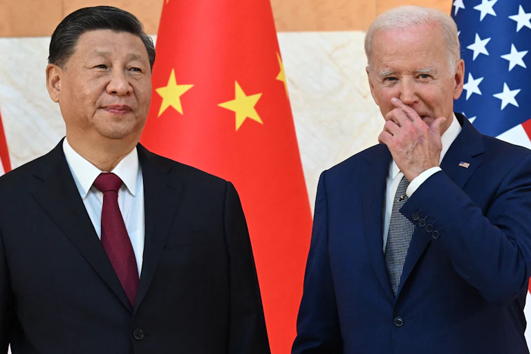 A man in a blue suit stands next to a man in a black suit. Both in front of flags.