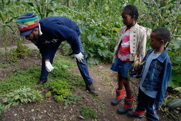 A man in a hat is gardening with two young children in boots.
