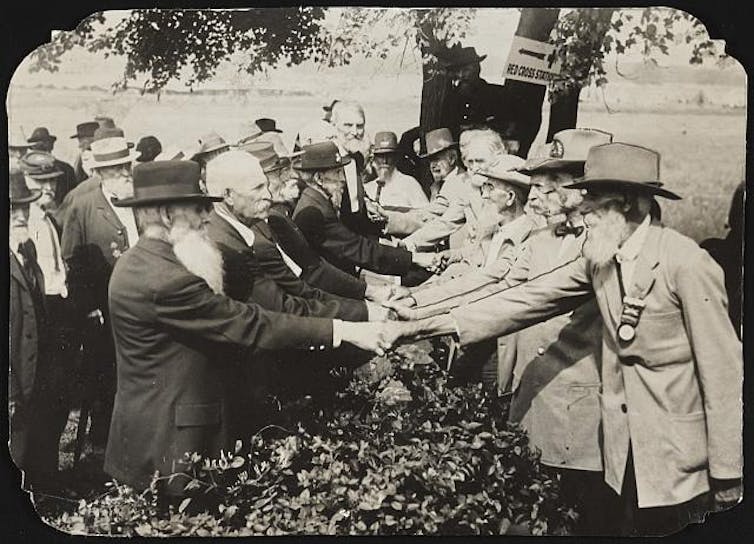 A black and white photo shows two rows of elderly men in suits -- one row in black, the other in light-colored fabric -- shaking hands.
