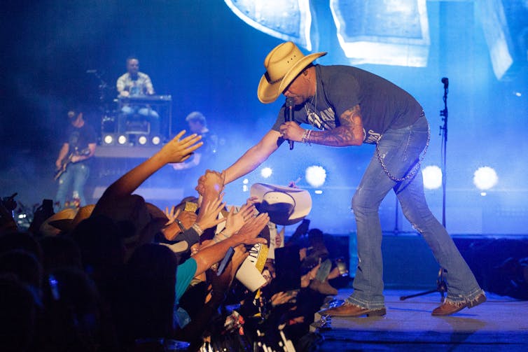 A white man wearing a cowboy hat shakes hands with spectators as he performs on stage.