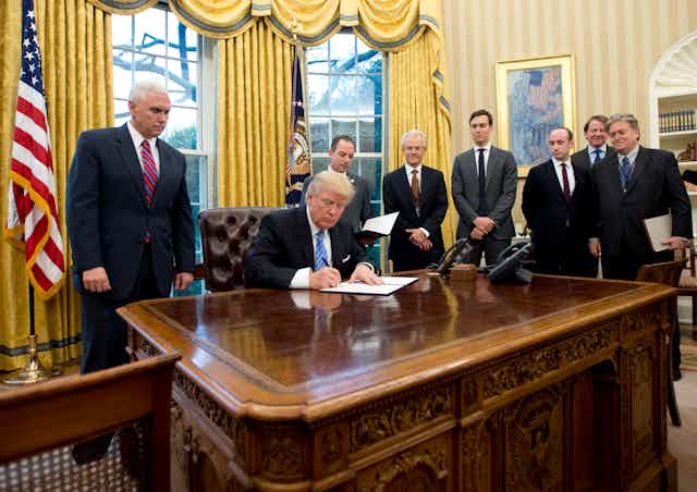 Seven men stand behind a man at a desk who is signing something in a fancy office.
