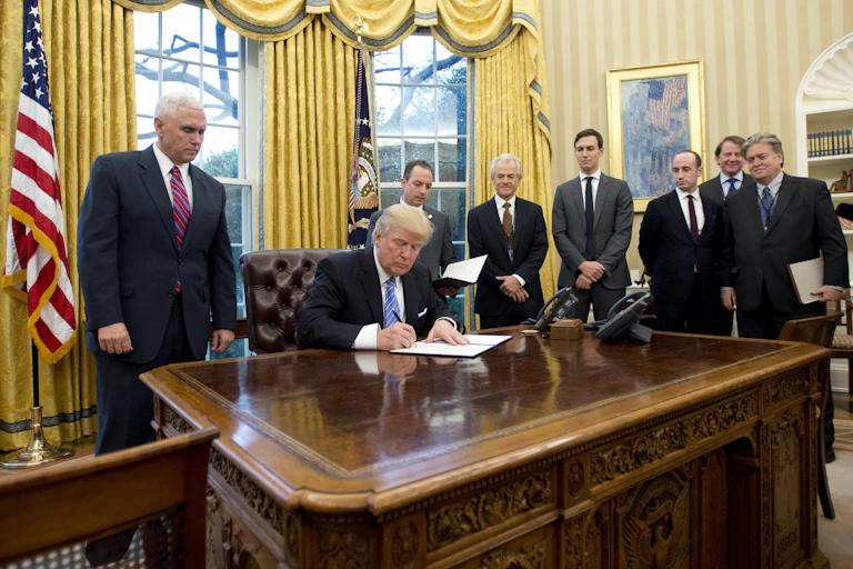 Seven men stand behind a man at a desk who is signing something in a fancy office.