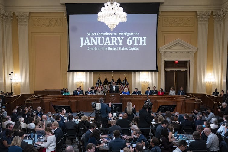 A large room filled with people, including almost a dozen at a long table at the front of the room, with a large screen behind them that says them that says 'Select Committee to Investigate the January 6th Attack on the U.S. Capitol.'