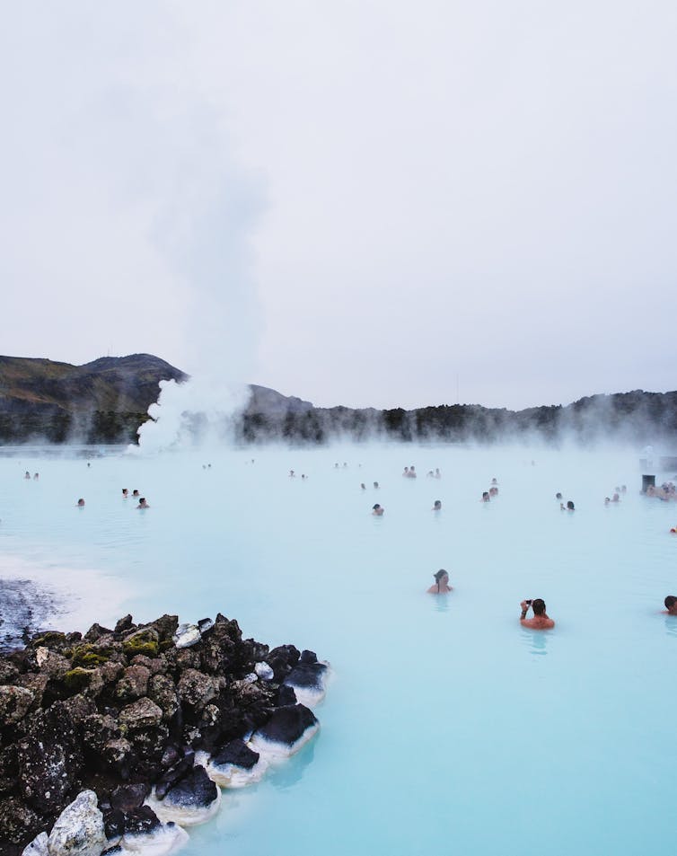 People sit in an eggshell-blue lake surrounded by black lava rocks. Steam rises in the background.