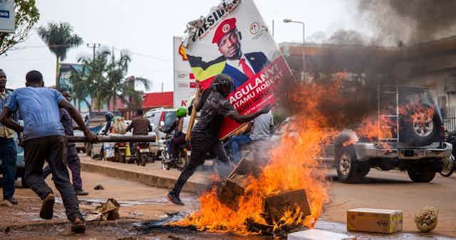 A man running past burning cartons while another is holding up a campaign poster on the side of a busy street