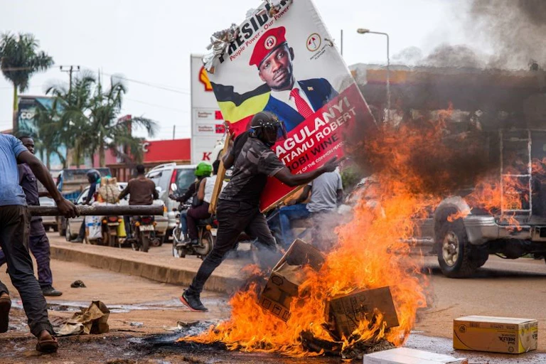 A man running past burning cartons while another is holding up a campaign poster on the side of a busy street
