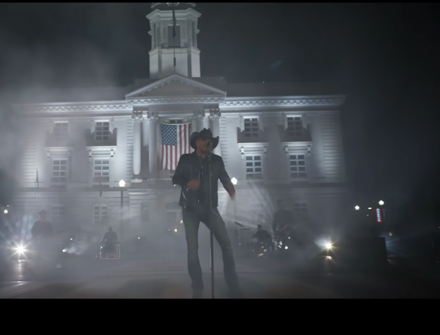 A man dressed in black holds a microphone as he sings in front of a courthouse that has an American flag in front of its windows.