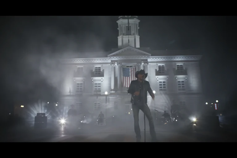 A man dressed in black holds a microphone as he sings in front of a courthouse that has an American flag in front of its windows.
