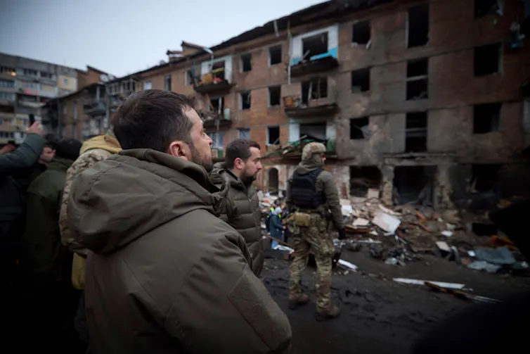 Ukraine's president Volodymyr Zelensky wearing a jacket looks at destroyed buildings.
