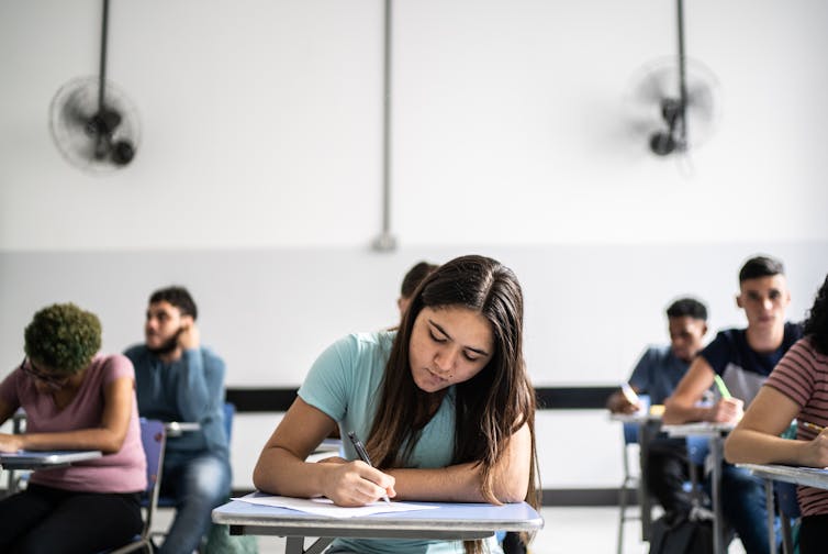 Students hunch over desks, writing on paper.