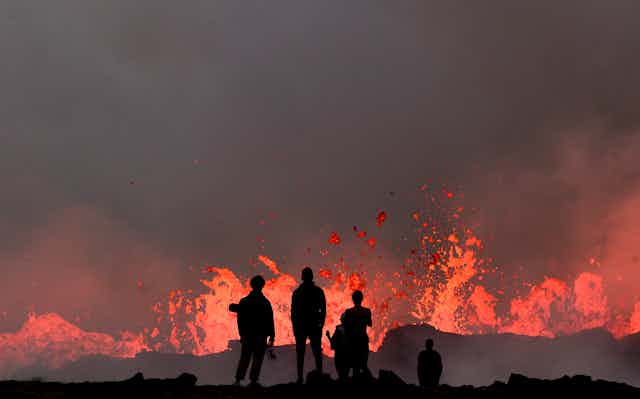 Several people watch a volcano's low-level eruption. The people are silhouetted against the lava.