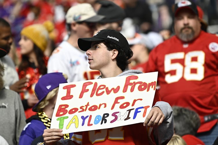 A football fan wearing a Chiefs jersey holds a handmade sign that says 