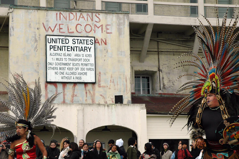Several people, some in traditional Native American attire, gather outside a building whose sign says - United States Penitentiary.