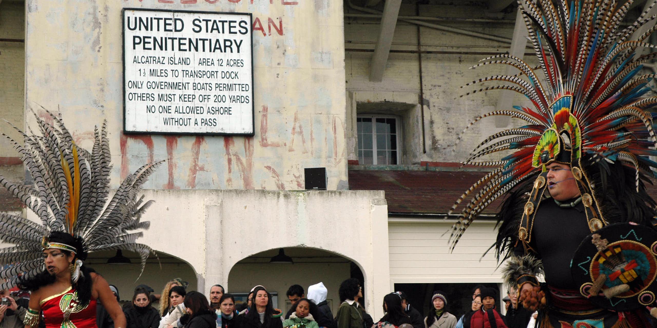 Several people, some in traditional Native American attire, gather outside a building whose sign says - United States Penitentiary.