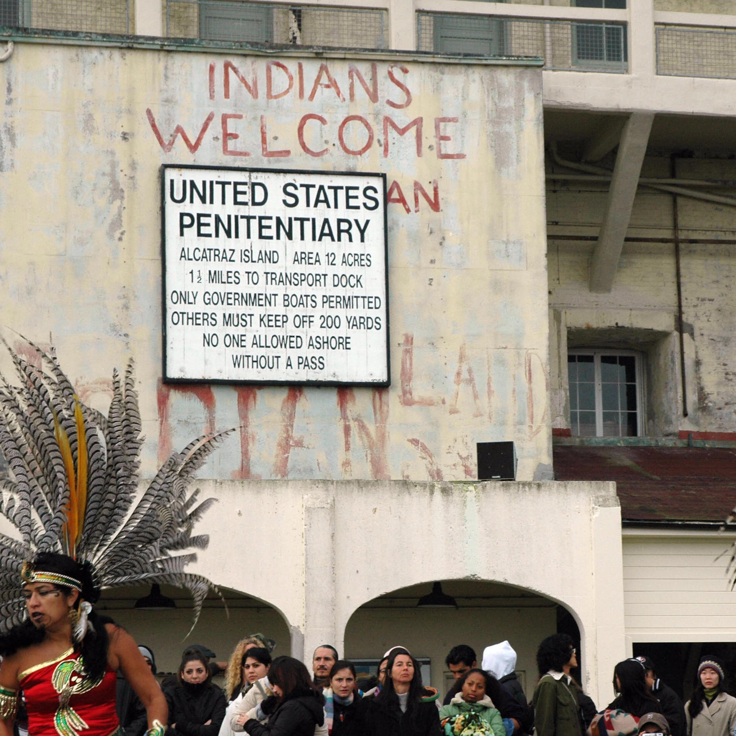 Several people, some in traditional Native American attire, gather outside a building whose sign says - United States Penitentiary.