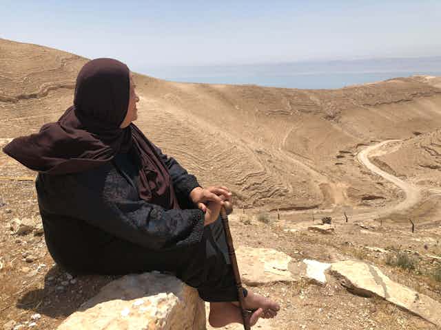 A woman sits on a wall and looks over a desert that leads to the sea.