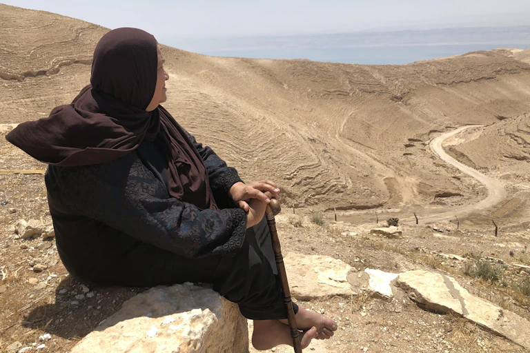 A woman sits on a wall and looks over a desert that leads to the sea.