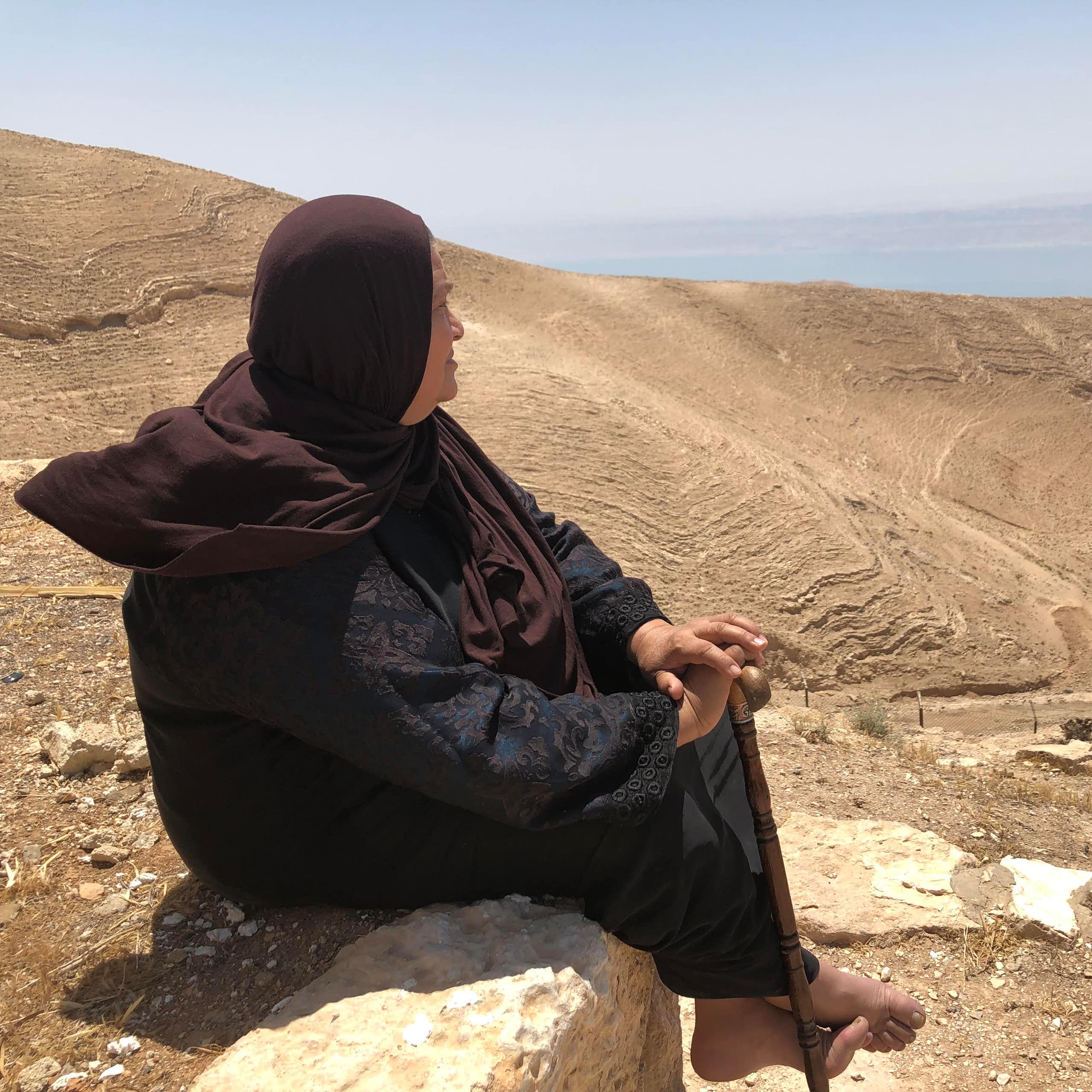 A woman sits on a wall and looks over a desert that leads to the sea.