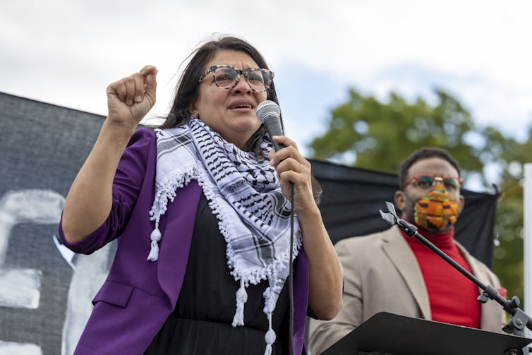 A woman in a black and white scarf speaks into a microphone