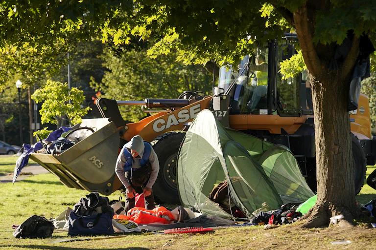 A man clearing out his belongings next to a tent, with a bucket loader behind him.