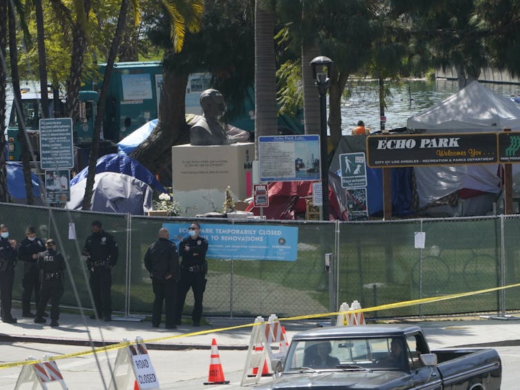 Police standing next to a chain-link fence around a park with tents in it.