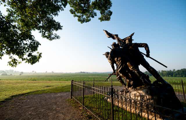 A statue of men holding rifles and a flag, positioned at an angle as though they are rushing into battle, is encircled by a low iron fence on a green field.