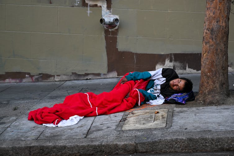 A dark-haired man sleeping in a red sleeping bag on a sidewalk.