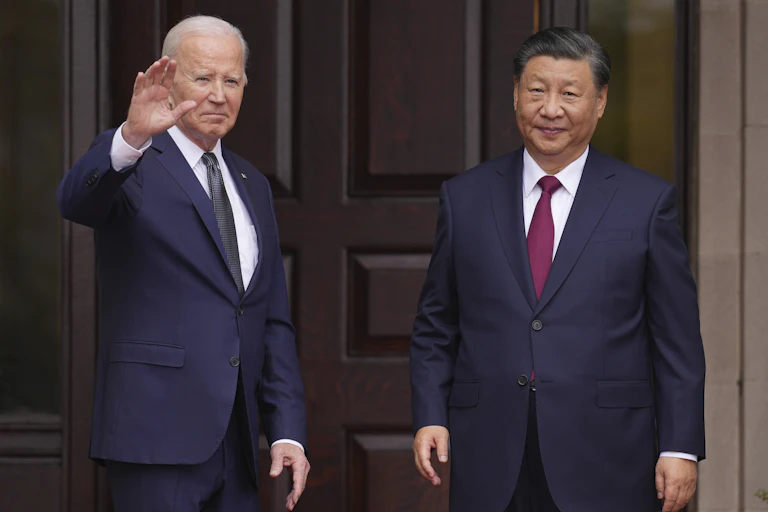 Two men in dark-blue suits stand in the entranceway of an estate in front of dark wood doors. The older man waves.