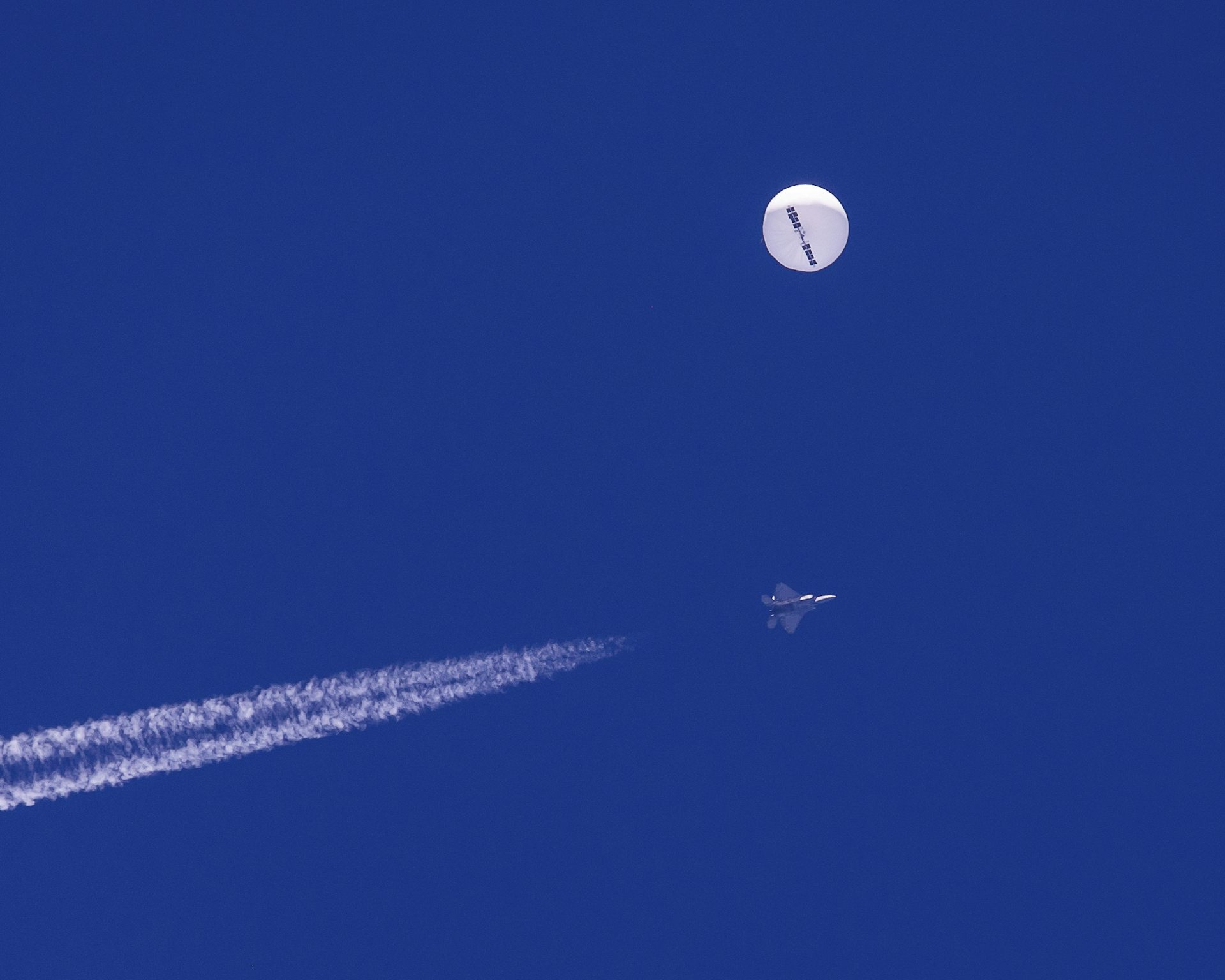 A bright round large balloon is seen in a blue sky with a fighter jet underneath it.