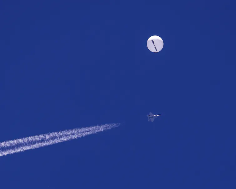 A bright round large balloon is seen in a blue sky with a fighter jet underneath it.