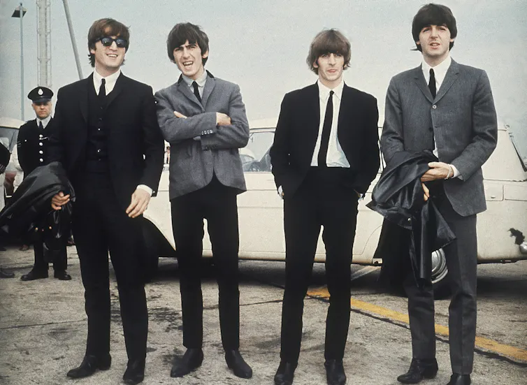 Four men seen in slim suits in front of a white car.