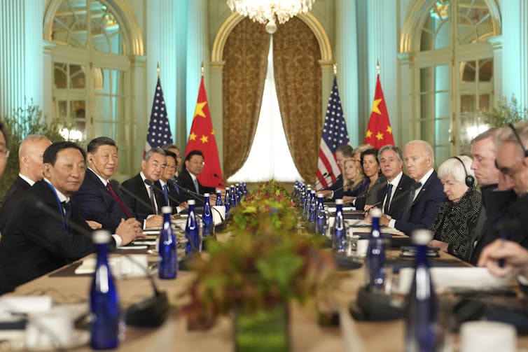 A group of men sit on either side of a long table. Flags are at the end of the table, and a large window with lush drapery.