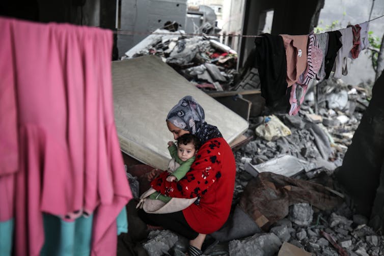A woman with a headscarf holds a small baby and sits amid a large pile of rubble.