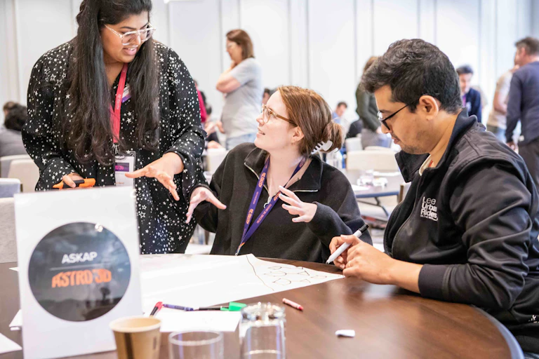 A photo of two women and a man talking at a table with some pens and paper on it. In front of them is a sign reading ASKAP ASTRO 3D.