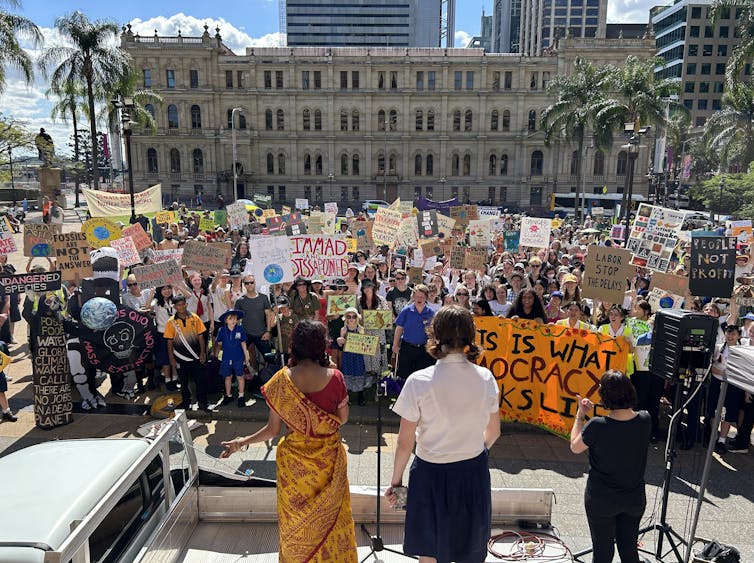 Australian school students and others striking & organizing to demand real action on the climate crisis in Brisbane, Australia. The crowd holding many signs and banners watches as two female speakers address the protestors in solidarity.