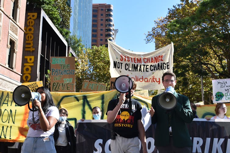 Image of a student strike for climate in Sydney, Australia in 2022. Students marching, three shown with megaphones, outside in organized protest with tall buildings and trees in the background.