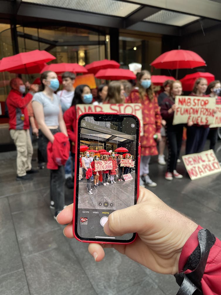 An image of a person's hand holding an iPhone while its camera captures a simultaneous image of school strikers protesting against fossil fuels outside of the National Australia Bank in December of 2021. Protestors were wearing medical masks and carrying red umbrellas, holding signs that generally say "stop funding fossil fuels".