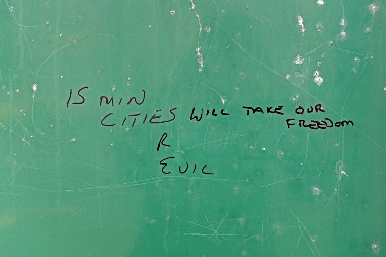 a green wall with handwritten graffiti saying 15 MINUTE CITIES R EVIL WILL TAKE OUR FREEDOM
