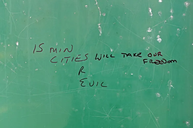 a green wall with handwritten graffiti saying 15 MINUTE CITIES R EVIL WILL TAKE OUR FREEDOM