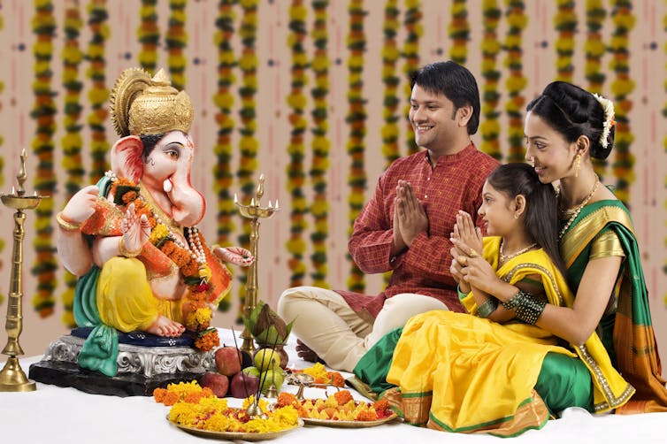A couple, with a young child in the woman's lap, sitting before the Hindu God Ganesha, with folded hands.