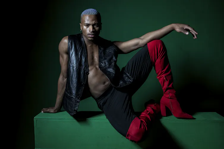 A man in a leather waistcoat and no shirt sits with a leg up and his arm draped over it.