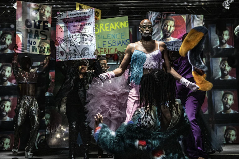 A stage set with a backdrop of posters and images of a man. A colourfully-dressed group of dancers perform.