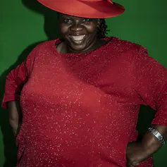 A woman smiles, hands on her hips in a shimmering red gown and hat.