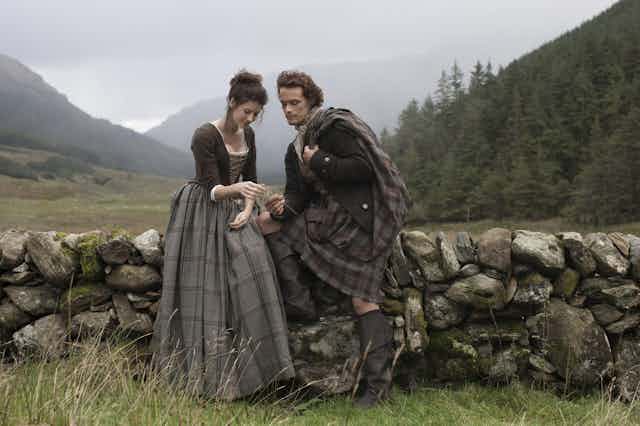 A couple in 18th-century Scots dress sitting on a dry stane dyke talking.