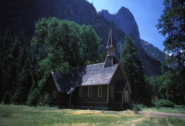 A small wooden church with a steeple stands against trees, with tall mountains in the background.