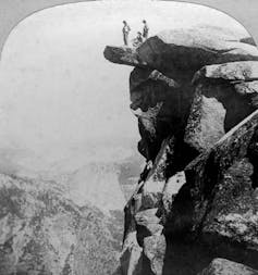 A black and white photo shows two men standing on a tiny ledge above a deep valley.