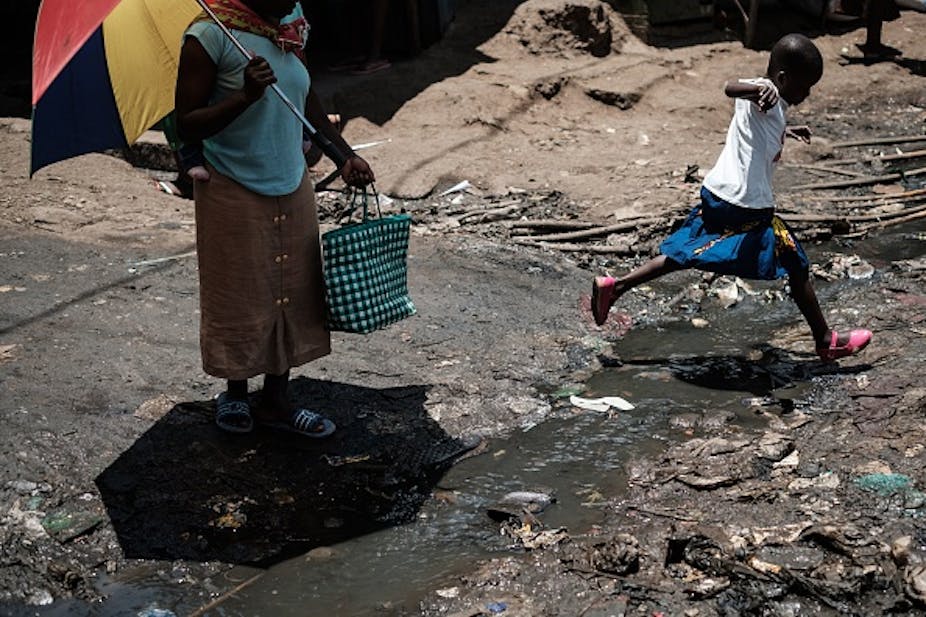 A child jumps over a stream of dirty water while an adult stands nearby.