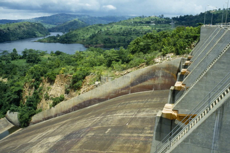 Concrete dam wall in the foreground, wooded cliffs and water in the background.